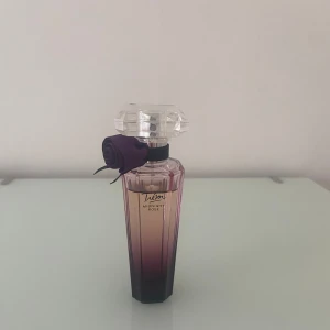 Trésor Midnight Rose Eau de Parfum - En elegant parfym från Lancôme med en lila-rosa glasflaska och dekorativ ros vid halsen. Doften har fruktiga och blommiga toner av hallon och ros, med inslag av svarta vinbär, rosépeppar, pion, jasmin, vanilj, mysk och cederträ.
