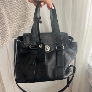 Svart handväska från Marc by Marc Jacobs - Säljer en svart handväska i skinn från Marc by Marc Jacobs. Väskan har silverfärgade metalldetaljer, dubbla handtag och en avtagbar axelrem. Klassisk design med lock och vridspänne framtill. Perfekt för dig som gillar stilrena accessoarer.