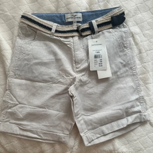 Pojkshorts från Hampton Republic – helt nya med etikett – storlek 146 - Stilrena pojkshorts från Hampton Republic i ljusbeige färg med flätat bälte. Helt nya med prislapp kvar. Storlek 146 (ca 10–11 år). Perfekta till vardags eller finare sommaranvändning.