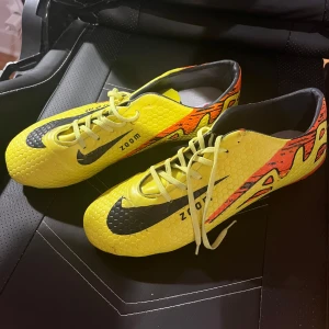 Gula  Zoom fotbollsskor - Snygga fotbollsskor  i en riktigt poppig gul färg med svarta detaljer och orange mönster på sidorna. Skorna har en texturerad yta med hexagonmönster, snörning och låg profil. Perfekta för dig som vill sticka ut på planen.