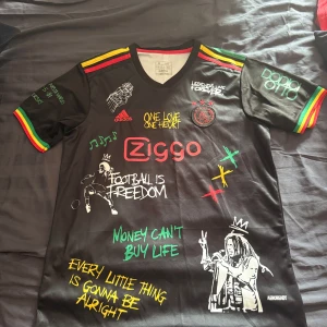 Ajax x Bob Marley Adidas fotbollströja - Unik svart Ajax fotbollströja från Adidas med Bob Marley-tema. Färgglada ränder i rött, gult och grönt på ärmar och axlar, tryck med texter och illustrationer på både fram- och baksida. V-ringad krage och tillverkad i lätt, ventilerande material.