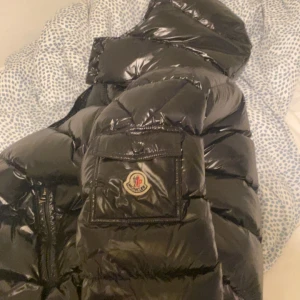 Svart Moncler dunjacka med huva - Säljer en svart, glansig dunjacka från Moncler med huva och klassisk logga på bröstfickan. Jackan har dragkedja och stora quiltade partier som ger en riktigt fet look. Perfekt för dig som vill ha en varm och snygg vinterjacka med streetkänsla.