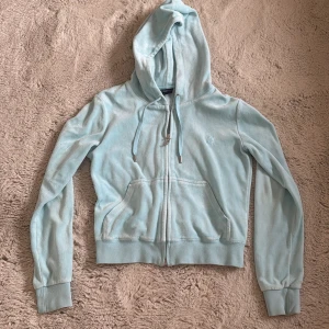 Ljusblå velour hoodie Juicy Couture - Supermjuk ljusblå hoodie från Juicy Couture i velour med dragkedja och huva. Klassisk J-detalj på dragkedjan och kängurufickor framtill. Perfekt för en chill och trendig look. Långa ärmar och ribbade muddar för extra komfort. Matrialet är lite som handuksmaterial.