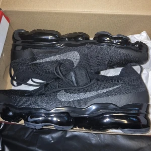 Nike Vapormax svarta sneakers - Svarta Nike Vapormax sneakers med stickad ovandel och silvrig swoosh på sidorna. Skorna har en unik, genomskinlig och bubblig sula som ger en futuristisk vibe. Perfekta för dig som gillar streetstyle och vill ha något som sticker ut.