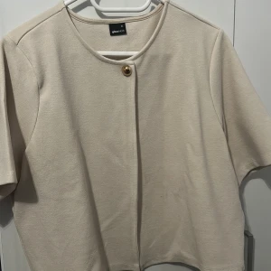 Beige cardigan jacka från Gina Tricot - Stilren beige cardigan jacka från Gina Tricot i storlek XL. Jackan har korta ärmar, rund halsringning och en snygg guldig knapp framtill. Enkel att matcha med olika outfits! Dock är knappen lite missfärgad och har tappat färg men det är inget man märker💕