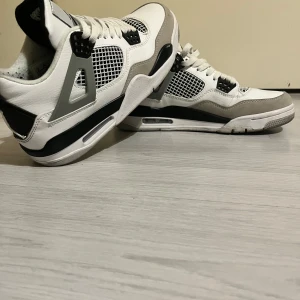 Jordans  4 - Nike Air Jordan 4 sneakers i vitt skinn med svarta och grå detaljer. Klassisk meshpanel på sidorna, Jumpman-logga på plös och häl, samt snörning framtill. Snygg kontrast mellan svart, grått och vitt gör dem till ett statement på fötterna.