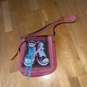 Rosa axelväska med sneakersdetalj - Cool rosa axelväska i bomull med justerbar rem och unik detalj framtill – två sneakers med riktiga skosnören i blått och vitt. Väskan har dragkedja och flera fack, samt kontrasterande bruna detaljer. Perfekt för dig som vill sticka ut med din stil.