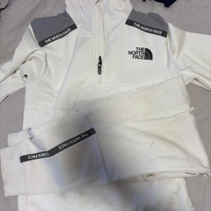 Vit half zip hoodie från The North Face - Säljer en vit hoodie från The North Face med grå axelpartier och svart logga på bröstet. Tröjan har half zip-dragkedja, huva och svarta detaljer med logotyp på ärmar och fickor. Materialet är mjukt och strukturerat, perfekt för kyliga dagar.