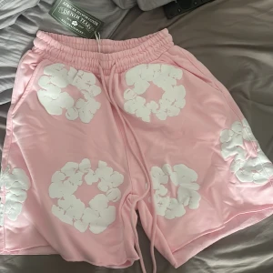 Rosa blommiga shorts  - Säljer ett par riktigt snygga rosa shorts  med vita blommiga mönster över hela plagget. De har elastisk midja med snörning och en bakficka. Perfekta för sommaren och ger en chill vibe till din outfit.
