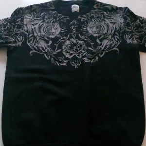 Svart tröja med tigrar från Zara - Svart tröja från Zara med grått mönster av tigrar och blommor över bröstet och axlarna. Tröjan har rund hals och korta ärmar, perfekt för dig som vill sticka ut med en cool och unik design.