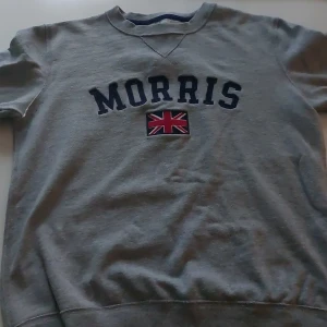 Grå sweatshirt från Morris XS - Grå sweatshirt från Morris i storlek XS med klassisk rund halsringning. Framsidan har tryckt Morris-logga och brittisk flagga i rött, vitt och blått. Tröjan är i mjuk bomull och har långa ärmar. Perfekt för en chill och stilren look.