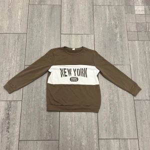 Brun långärmad tröja New York Shein - Brun långärmad tröja från Shein med vit panel över bröstet och trycket 'NEW YORK 1995'. Tröjan har rund halsringning och normal passform. Perfekt för dig som gillar streetstyle och enkel design.