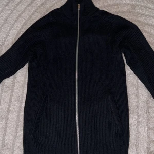 Svart stickad kofta från Jack & Jones - Svart ribbstickad kofta från Jack & Jones med hel dragkedja framtill och hög krage. Tröjan har långa ärmar och två fickor på sidorna. Perfekt för lager-på-lager och enkel att matcha med olika outfits. Storlek S, passar även M!😊