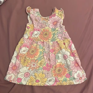 Blommig kortklänning från H&M, strl 80 - Supersöt kortklänning från H&M i storlek 80 med färgglatt blommönster i rosa, gul, orange och vitt. Klänningen har volangdetaljer vid axlarna och är ärmlös, perfekt för varma dagar. Tillverkad i mjuk bomull som känns skön mot huden.