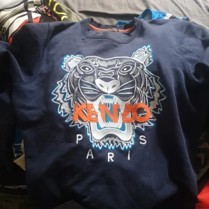 Mörkblå Kenzo sweatshirt med tiger - Säljer en mörkblå sweatshirt från Kenzo med ikoniskt broderat tigermotiv och orange Kenzo-text på bröstet. Tröjan har rund halsringning, ribbade muddar och är i mjuk bomull. Perfekt för dig som vill sticka ut med en streetstyle-look.