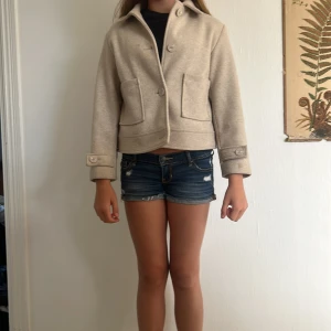 Beige kort ulljacka med stora knappar - Kort beige jacka i ull med stora knappar framtill och på ärmsluten. Jackan har två stora fickor framtill och en klassisk krage. Snygg loose passform som ger en chill vibe och passar perfekt till höst eller vår.