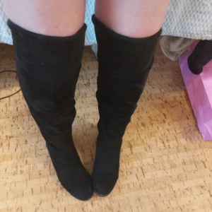Svarta höga mockastövlar - Snygga svarta stövlar i mocka med högt skaft som går över knäet. De har en klassisk rund tå och platt sula, vilket gör dem både stilrena och bekväma. Perfekta att matcha med kjol eller klänning för en trendig look.