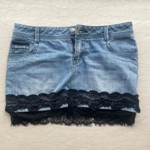 Jeanskjol med svart spetskant - Snygg kort jeanskjol i ljusblå denim med bred svart spetskant nertill. Klassisk femficksmodell med bälteshällor och dragkedja fram. Spetsen ger kjolen en extra cool och feminin touch. Perfekt för dig som vill sticka ut lite extra.