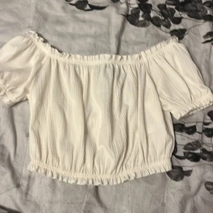Offshoulder vit blus med puffärm - Superfin offshoulder blus i vitt med korta puffärmar och smockdetaljer vid ärmslut och midja. Tunt, krinklat tyg som ger en luftig känsla och snygg struktur. Perfekt till jeans eller kjol för en fräsch look.
