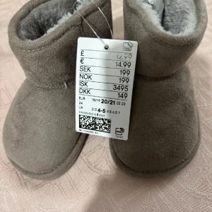 Gråa mjuka babyboots i mocka - Supersöta gråa babyboots med mjukt foder och utsida i mocka. Perfekta för att hålla små fötter varma och mysiga under kyliga dagar. De har en enkel och stilren design med rund tå och platt sula.