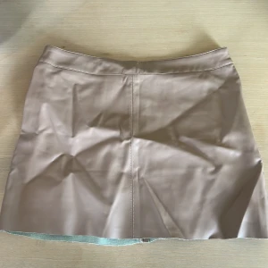 Beige kort kjol i skinnimitation - Snygg beige kortkjol i skinnimitation med stilren design och raka sömmar. Perfekt för dig som vill ha en trendig och clean look. Kjolen har en enkel passform och är lätt att matcha med olika toppar och jackor.