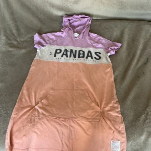 LC Waikiki Pandas hoodieklänning - Superchill hoodieklänning från LC Waikiki i lila, vit och ljusrosa. Stor tryckt text 'PANDAS' framtill, stor ficka och korta ärmar. Perfekt för dig som gillar avslappnad stil och gulliga detaljer.