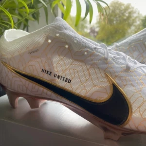 Nike Zoom Vapor 15 Elite FG fotbollsskor - Oanvända och kvitto finns!Nike Zoom Vapor 15 Elite FG i vitt med guld- och svarta detaljer. Ovandel i Flyknit-material med snyggt geometriskt mönster och stor svart Swoosh. Yttersula i metallic roséguld med Zoom Air-dämpning. Perfekta för dig som vill sticka ut på planen. 