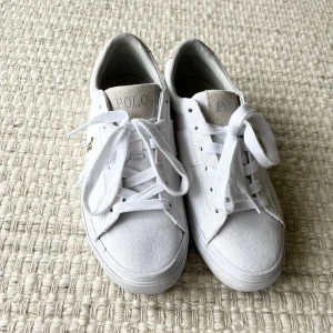 Vita sneakers från Polo Ralph Lauren - Fräscha vita sneakers från Polo Ralph Lauren med klassisk design. Skorna har canvasmaterial, snörning och detaljer i beige mocka på häl och plös. Broderad Polo-logga på sidan och tydlig POLO-text bak. Perfekta för en clean och stilren look.