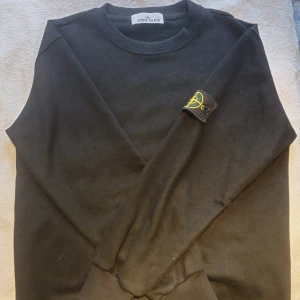 Svart Stone Island sweatshirt - Svart sweatshirt från Stone Island med klassisk rund hals och ikoniskt gult och grönt logomärke på vänster ärm. Tröjan är i bomull och har en clean, stilren look som passar perfekt till streetwear. Långärmad och normal passform.
