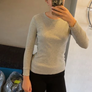 Grå långärmad tröja från H&M - En enkel och stilren grå långärmad tröja från H&M Basic, gjord i mjuk ekologisk bomull. Tröjan har rund halsringning och en slim passform som sitter snyggt på kroppen. Perfekt att matcha med jeans eller kjol för en clean look. 💕