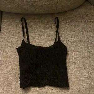 Svart linnetopp från Gina Tricot - Säljer en svart linnetopp från Gina Tricot med smala axelband och crinklad struktur. Toppen är croppad och har en enkel, stilren design som passar perfekt till jeans eller kjol. Superenkel att matcha och riktigt snygg till sommaren!