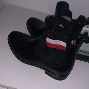 Svarta chelsea boots Tommy Hilfiger - Snygga svarta chelsea boots i glansigt syntetmaterial från Tommy Hilfiger. Resårpartiet har klassiska ränder i rött, vitt och blått samt en liten metallogo på sidan. Perfekta för en stilren och sportig look.