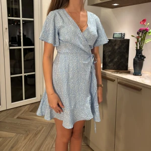 Ljusblå omlottklänning från SHEIN - Superfin ljusblå kort omlottklänning från SHEIN med vitt leopardmönster. Klänningen har v-ringning, volangkant nertill, korta vida ärmar och knyt i midjan. Perfekt för sommarens alla tillfällen och ger en fräsch och trendig look. Finns hål inuti klänningen där man trär bandet igenom.