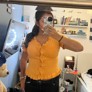 Gul ribbad blus med volangkant - Superfin gul ribbad blus med korta ärmar och v-ringning. Blusen har volangdetaljer längs knäppningen och nederkanten, vilket ger en söt och trendig look. Perfekt för dig som vill sticka ut med färg och stil i sommar.