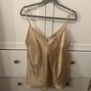Champagnefärgad linnetopp i satin - Säljer en stilren champagnefärgad linnetopp från H&M i glansigt satinmaterial. Toppen har smala axelband och v-ringning framtill, perfekt för en trendig och enkel look. Passar snyggt till både jeans och kjol. Den har varit älskad i många år🩷