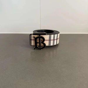 Rutig Burberry bälte med B-spänne - Snyggt bälte från Burberry med klassiskt rutigt mönster i beige, svart och rött. Bältet har ett stort svart B-spänne framtill och är gjort i skinn med tygdetaljer. Perfekt accessoar för att lyfta din outfit med en lyxig touch.