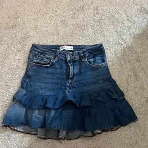 Jeanskjol med volang från Zara - Supersnygg jeanskjol från Zara i klassisk blå denim. Kjolen har volangdetalj nertill, fem fickor och bälteshällor. Stängs med dragkedja och knapp framtill💕