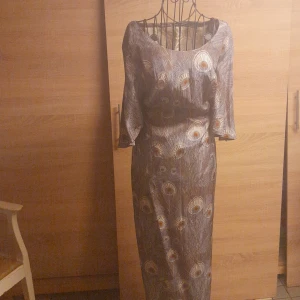 Långklänning med påfågelfjädermönster - Unik långklänning med gråbrun bas och mönster av påfågelfjädrar i beige och brunt. Klänningen har trekvartsärm och rund halsringning. Materialet har en lätt glansig yta som ger en elegant känsla. Dragkedja bak för enkel påklädning.