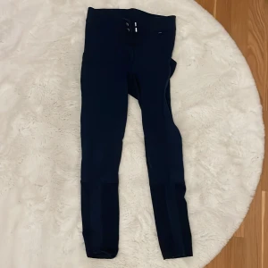 Mörkblå ridbyxor CRW, storlek 38 - Snygga mörkblå ridbyxor från CRW i storlek 38. Byxorna har hög midja, två silvriga knappar framtill och tight passform. Materialet är stretchigt och bekvämt, perfekt för ridning. Klassisk look med förstärkning på insidan av benen.