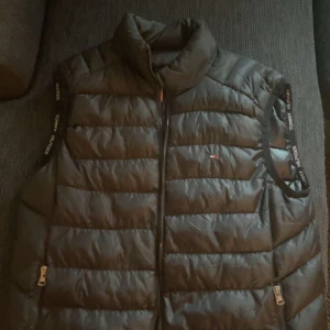 Svart dunväst från Tommy Hilfiger - Snygg svart dunväst från Tommy Hilfiger med quiltad design och dragkedja framtill. Västen har två fickor med dragkedja och logotypdetaljer på bröstet och insidan. Axelbanden har Tommy Hilfiger-tryck och insidan har en röd dragkedjeficka.