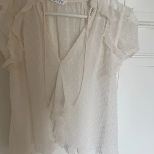 Vit blus med volang och knyt - Supersöt vit blus från Zara Basic med korta ärmar, volangdetaljer och knyt framtill. Tunt, lätt tyg med diskret prickigt mönster och v-ringning. Tröjan är väldigt genomskinlig kan behövas ett linne under 
