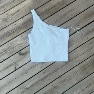 Ljusblå oneshoulder topp Gina Tricot - Säljer en ljusblå ribbad oneshoulder topp från Gina Tricot i storlek M. Toppen är ärmlös med en smal axelrem på ena sidan och har en croppad passform. Tillverkad i mjuk polyesterblandning med stretch för en bekväm känsla.