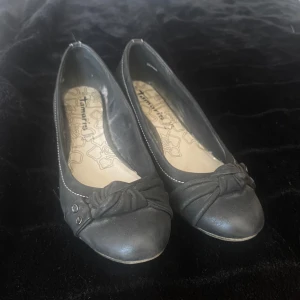 Svarta ballerinaskor med kilklack från Tamaris - Snygga svarta ballerinaskor från Tamaris i skinn med kilklack i trälook. Skorna har rund tå, dekorativ knut och nitar framtill samt vita sömmar längs kanten. Perfekta för dig som vill ha en klassisk men ändå trendig look.