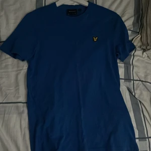 Blå t-shirt från Lyle & Scott OBS frakt tillkommer  - Snygg blå t-shirt från Lyle & Scott med klassisk rund hals och korta ärmar. På bröstet sitter den ikoniska gula örnloggan. T-shirten är i mjuk bomull och har en clean, stilren look som passar perfekt till jeans eller shorts. Kan mötas upp i centrala Göteborg eller står köparen för frakten 