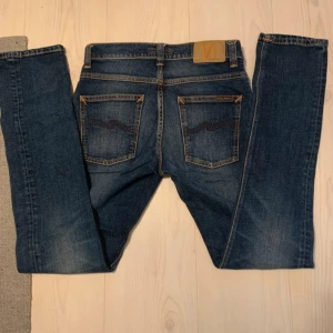 Nudie Jeans blå jeans W29 L32 - Säljer ett par klassiska blå jeans från Nudie Jeans med snygga slitningar och kontrastsömmar. Modellen har raka ben, fem fickor och coola detaljer på bakfickorna. Perfekta för dig som gillar en tidlös och avslappnad stil.