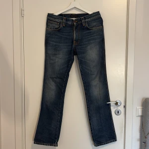 Blå jeans från Nudie Jeans, W32 L32 - Snygga blå jeans från Nudie Jeans med klassisk femficksdesign och orange kontrastsömmar. Jeansen har rak passform och normal midja, tillverkade i slitstarkt denim. Perfekta för dig som gillar en tidlös och avslappnad stil.