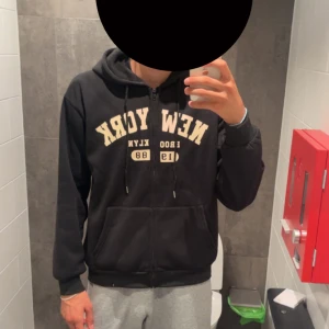 Svart hoodie med New York-tryck - Svart hoodie med dragkedja och huva, stora fickor framtill och snörning i halsen. Framsidan har ett beige New York-tryck och texten 'BROOKLYN 1988'. Perfekt för chill dagar eller när du vill ha en avslappnad streetstyle. Storlek L 