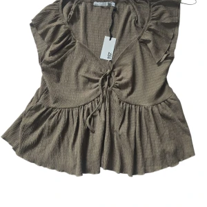 Brun ribbad blus med snörning XL - Supersöt brun blus från 157 i storlek XL. Toppen har ribbad struktur, volangdetaljer vid axlarna och en snygg snörning framtill. Den har v-ringning och är ärmlös, perfekt för en chill och trendig look.