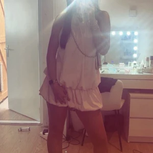 Champagnefärgad kort volangklänning - Supersöt kort klänning i champagnefärg med volanger och halterneck. Öppen rygg med knyt i nacken och luftig passform. Perfekt för sommarens alla tillfällen när du vill sticka ut lite extra. Materialet känns lätt och flowy.