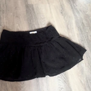 Svart prickig kortkjol Gina Tricot - Supersöt svart kortkjol från Gina Tricot med små glittriga prickar och bred, veckad midja. Kjolen är luftig och har en lätt volangig känsla, perfekt för dig som gillar en lekfull och trendig stil.